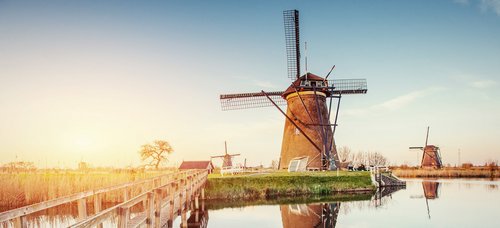 Traditionelle niederländische Windmühlen in einer grünen Landschaft mit blauem Himmel am Kanal von Rotterdam. Im Vordergrund eine schmale Holzbrücke über dem Kanal.