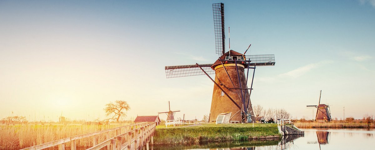 Traditionelle niederländische Windmühlen in einer grünen Landschaft mit blauem Himmel am Kanal von Rotterdam. Im Vordergrund eine schmale Holzbrücke über dem Kanal.